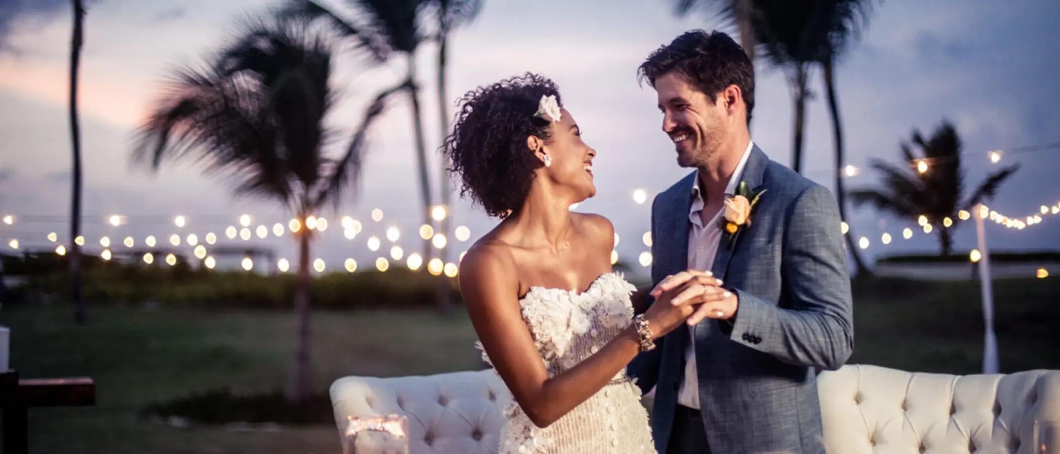 A happy bride and groom holding hands and smiling at each other during their wedding reception at sunset.