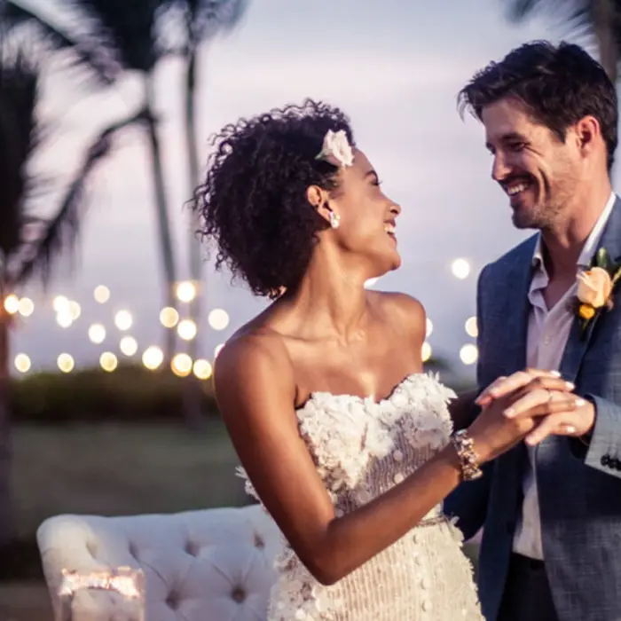 A happy bride and groom holding hands and smiling at each other during their wedding reception at sunset.