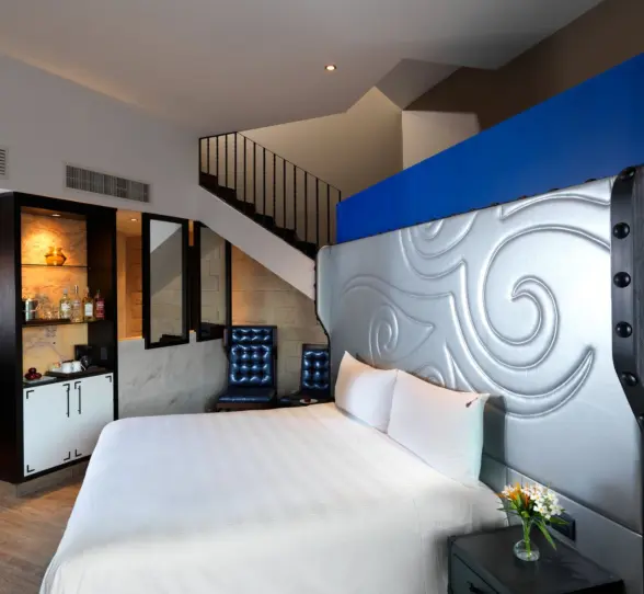 A modern, two-story hotel room with a white bed, blue accents, and a staircase leading to a sky terrace.