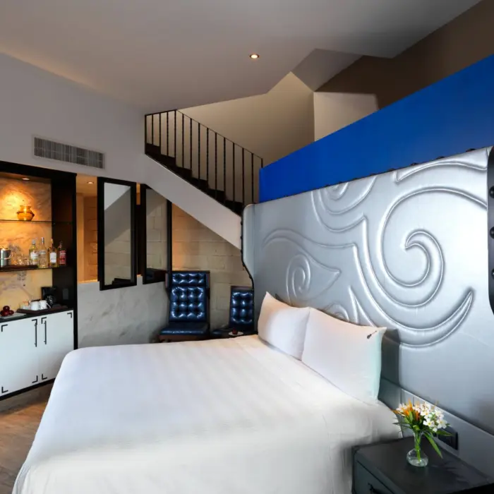 A modern, two-story hotel room with a white bed, blue accents, and a staircase leading to a sky terrace.