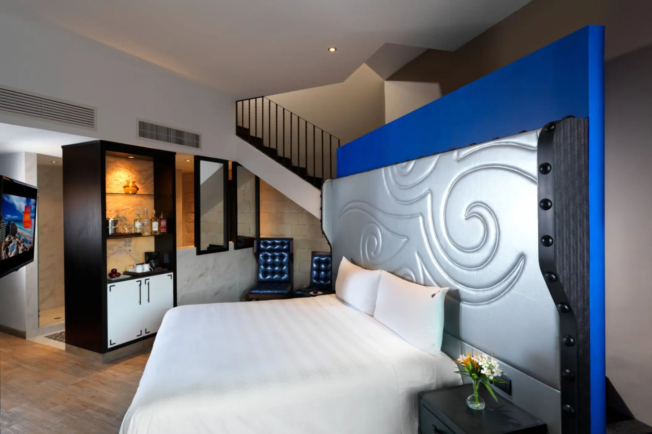 A modern, two-story hotel room with a white bed, blue accents, and a staircase leading to a sky terrace.