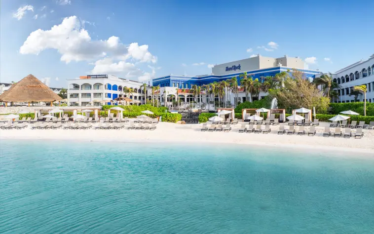 The Hard Rock Hotel Riviera Maya from the beach, showing the resort buildings and turquoise water.