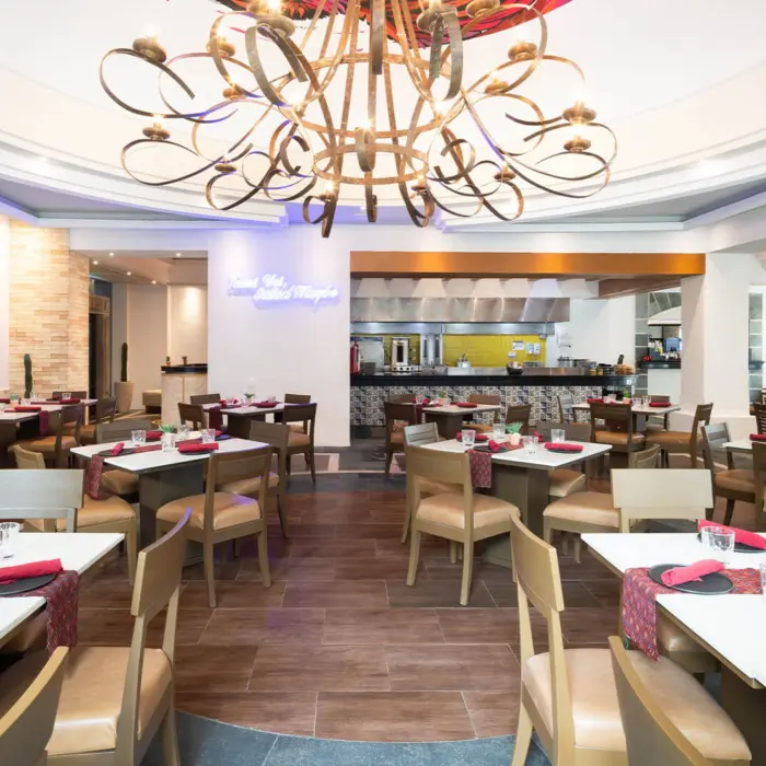 A brightly lit dining room with an open kitchen, formal table settings, and a large chandelier.