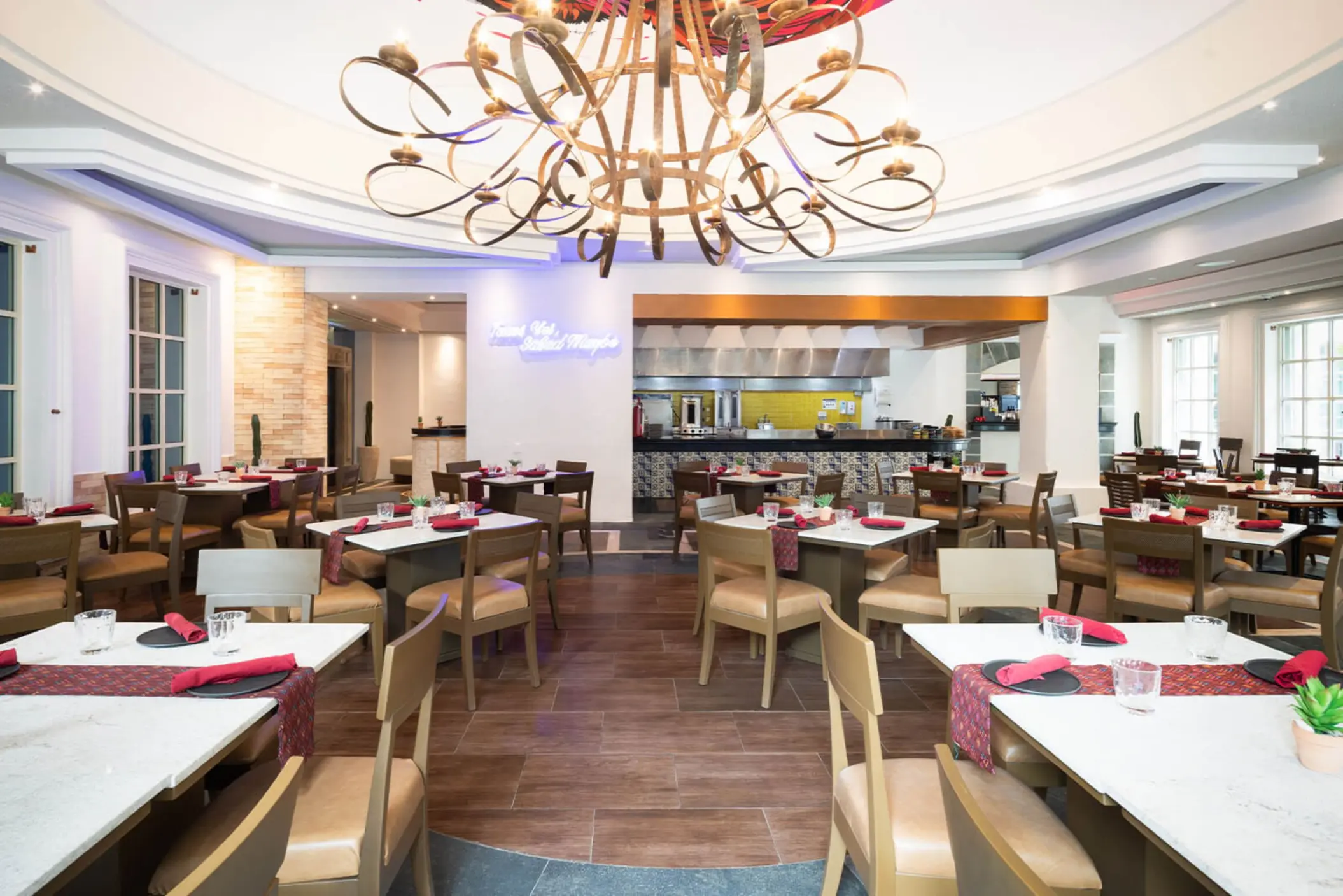 A brightly lit dining room with an open kitchen, formal table settings, and a large chandelier.
