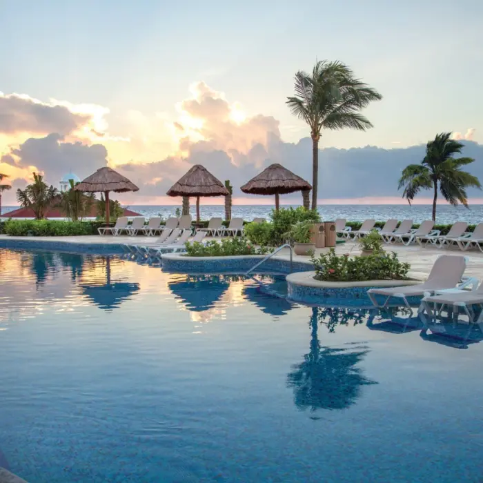 A tranquil view of a resort pool at sunrise with palm trees and lounge chairs.