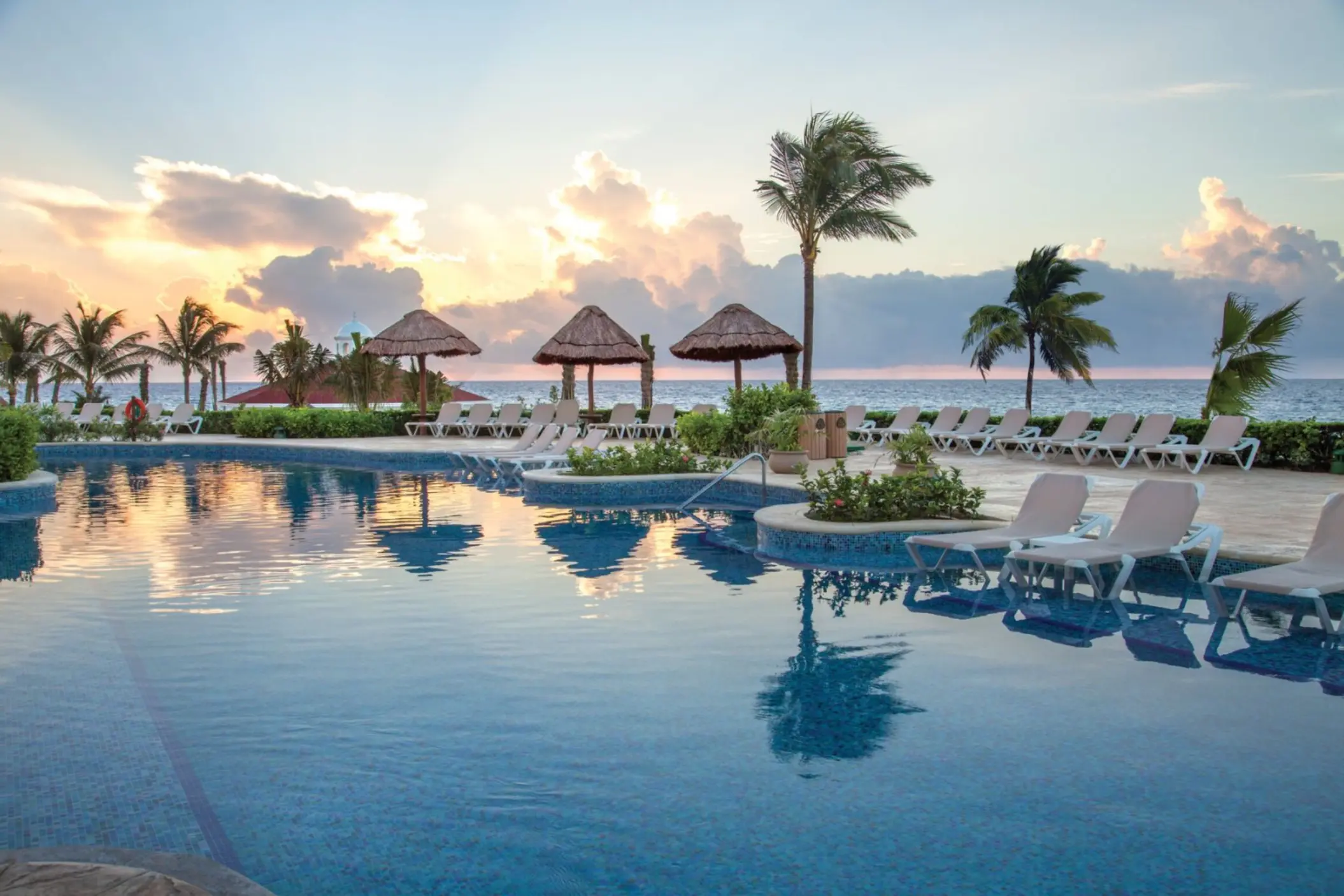 A tranquil view of a resort pool at sunrise with palm trees and lounge chairs.