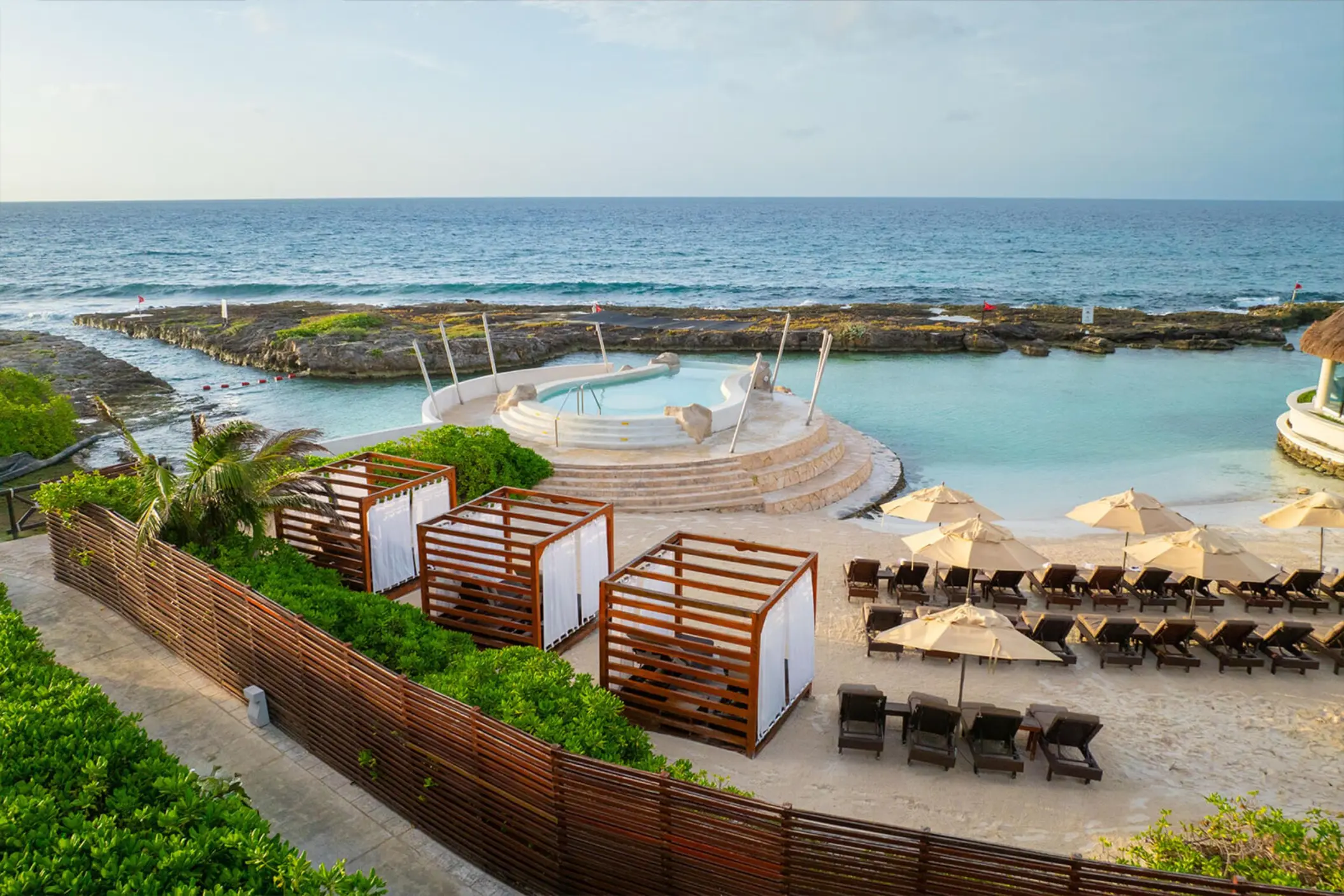 An elevated view of a protected cove beach with lounge chairs, cabanas, and a circular pool.