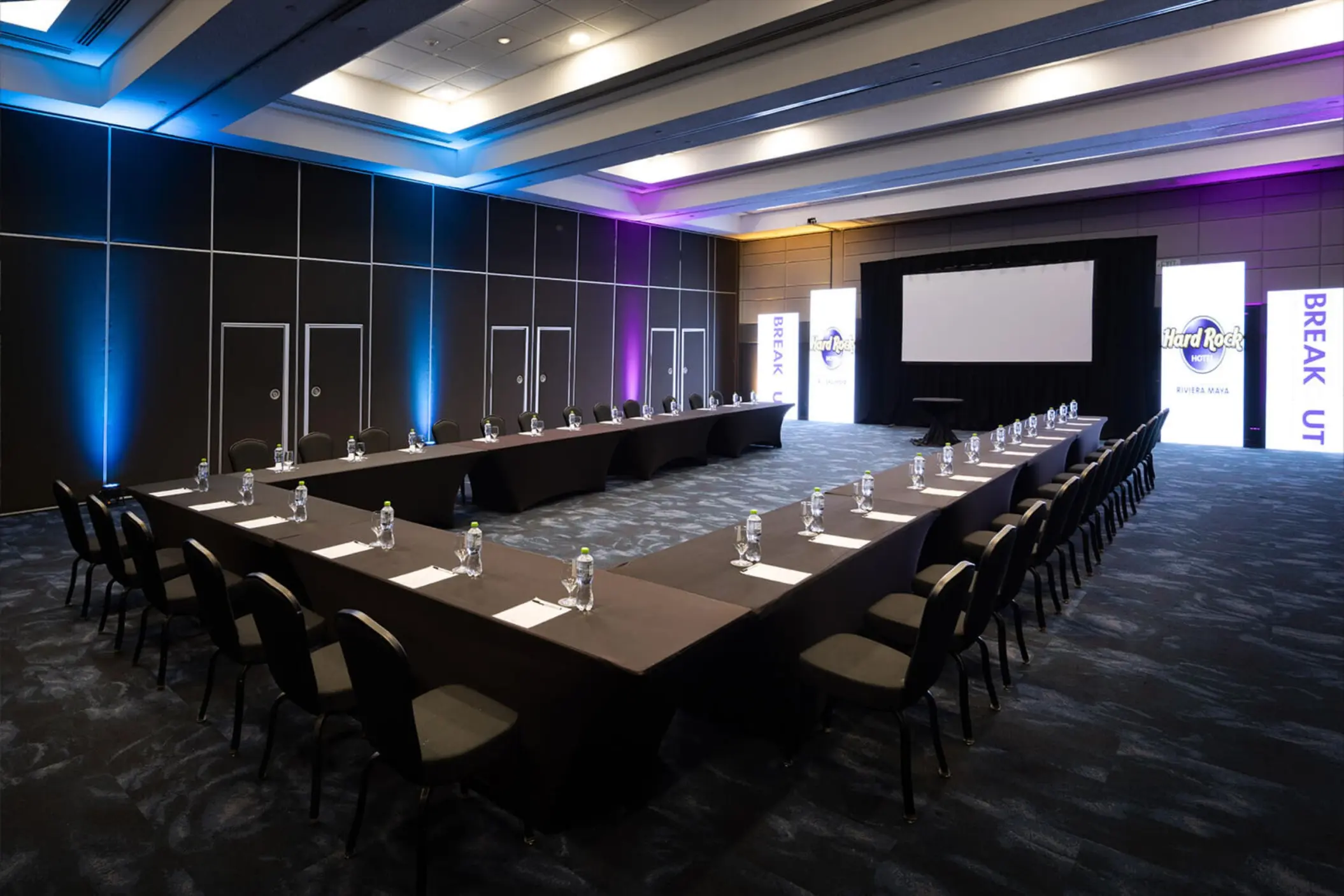 A modern U-shaped conference room with a large projector screen and purple and blue lighting.