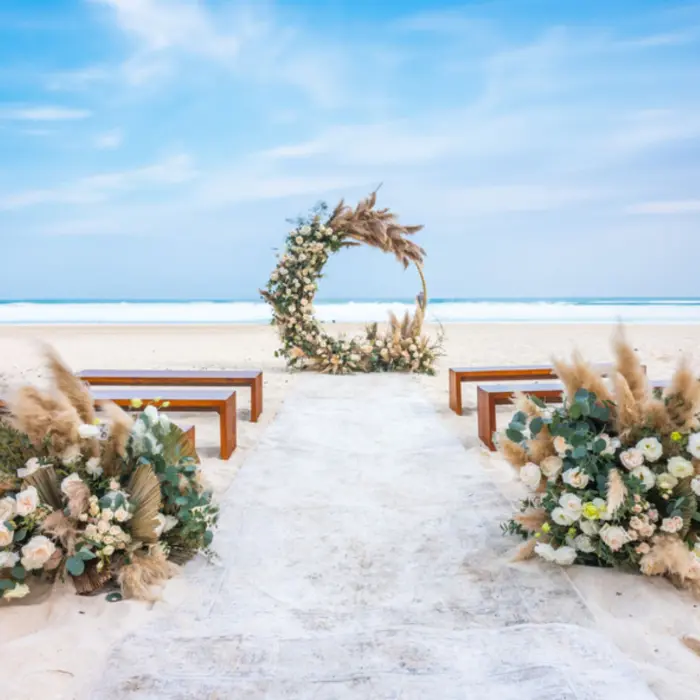 A beach wedding venue with a circular floral arch, wooden benches for guests, and a white aisle runner on the sand.