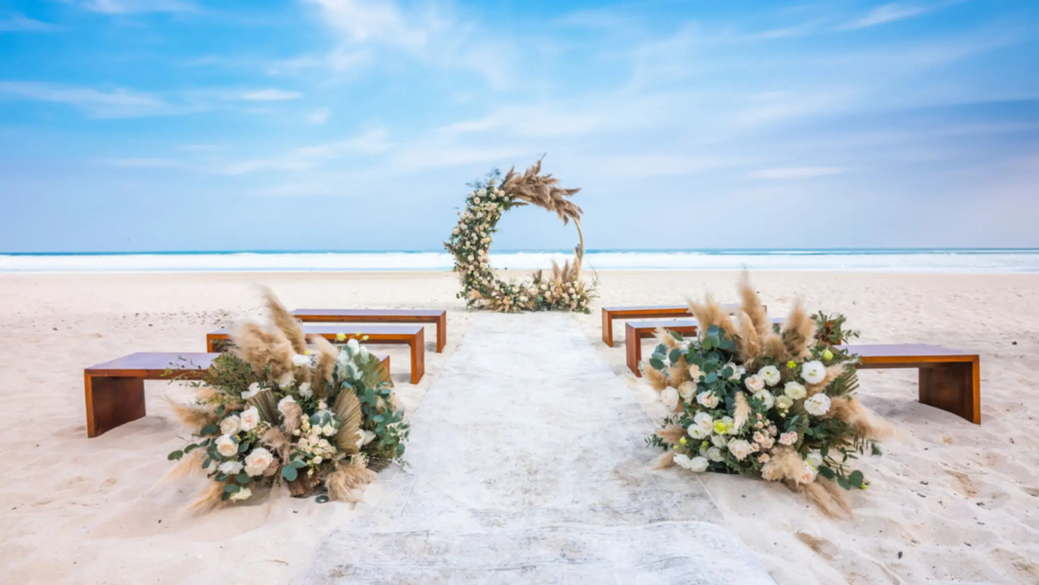 A beach wedding venue with a circular floral arch, wooden benches for guests, and a white aisle runner on the sand.