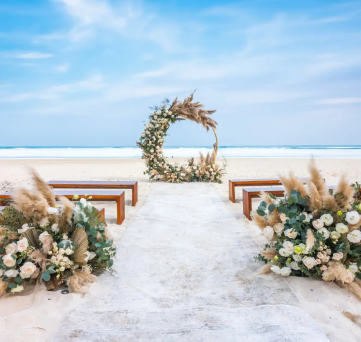 A beach wedding venue with a circular floral arch, wooden benches for guests, and a white aisle runner on the sand.