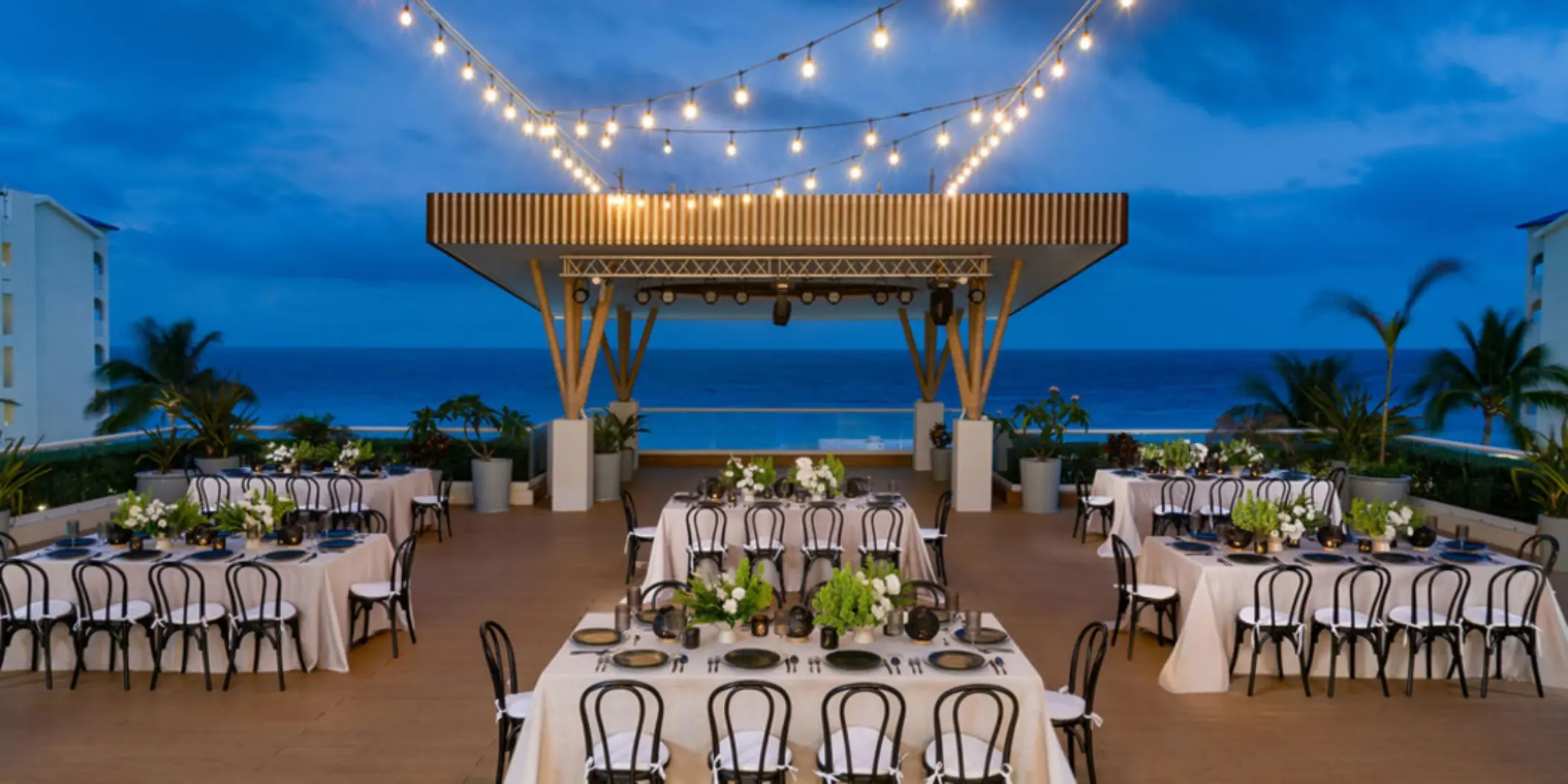 A rooftop event venue at dusk overlooking the ocean, with tables set for a banquet under a string of lights.