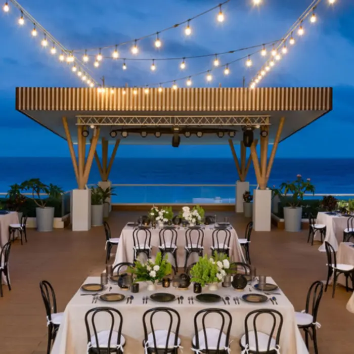 A rooftop event venue at dusk overlooking the ocean, with tables set for a banquet under a string of lights.