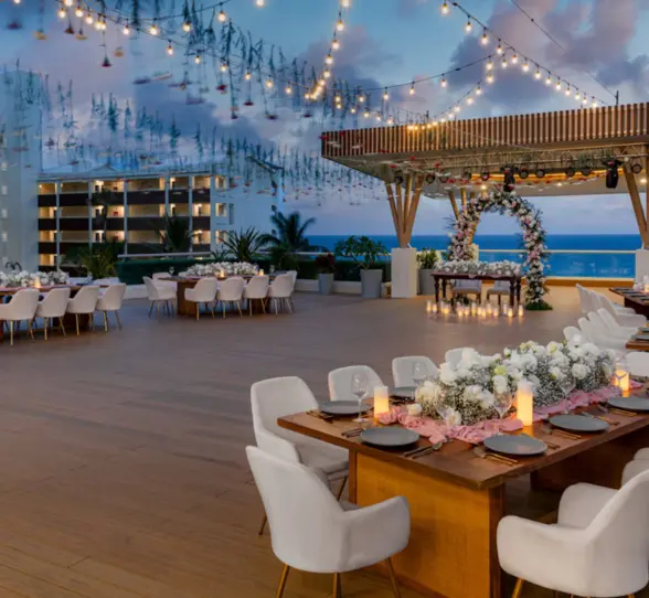 A beautiful outdoor rooftop dinner setting at dusk with string lights, floral centerpieces, and tables set for a banquet.
