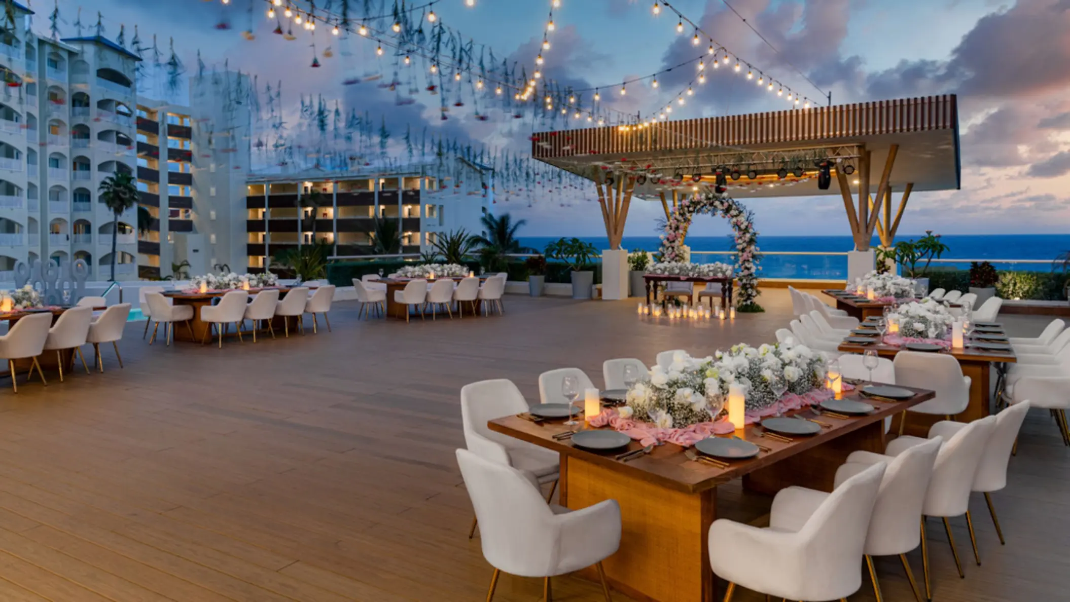 A beautiful outdoor rooftop dinner setting at dusk with string lights, floral centerpieces, and tables set for a banquet.
