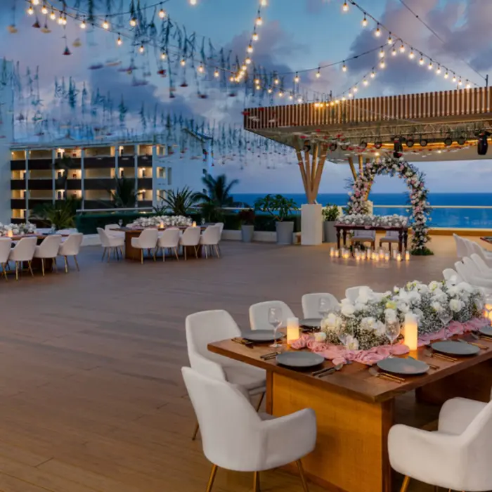 A beautiful outdoor rooftop dinner setting at dusk with string lights, floral centerpieces, and tables set for a banquet.