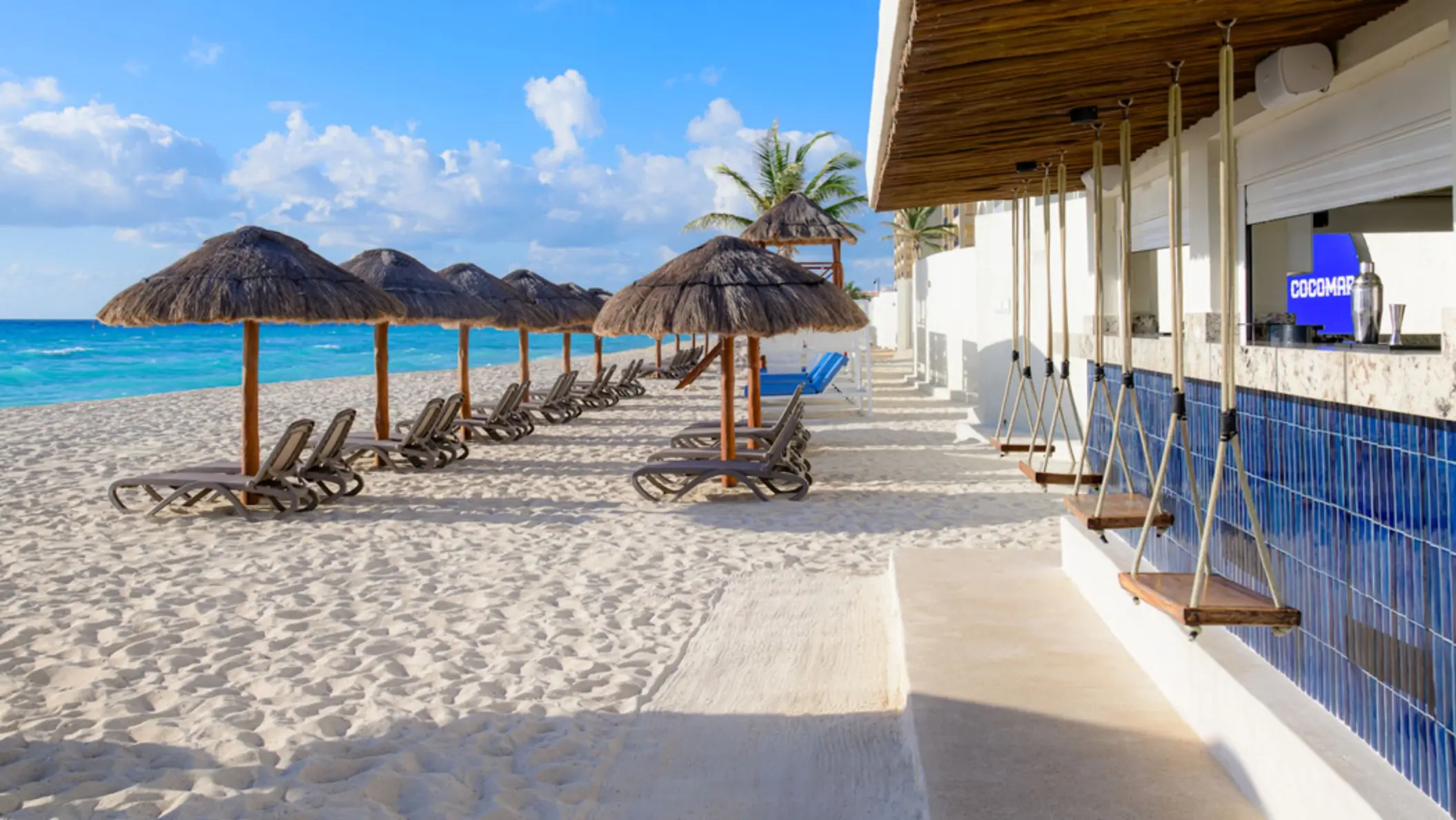A relaxing beach bar with thatched-roof umbrellas, lounge chairs on white sand, and swing seats at the bar counter.