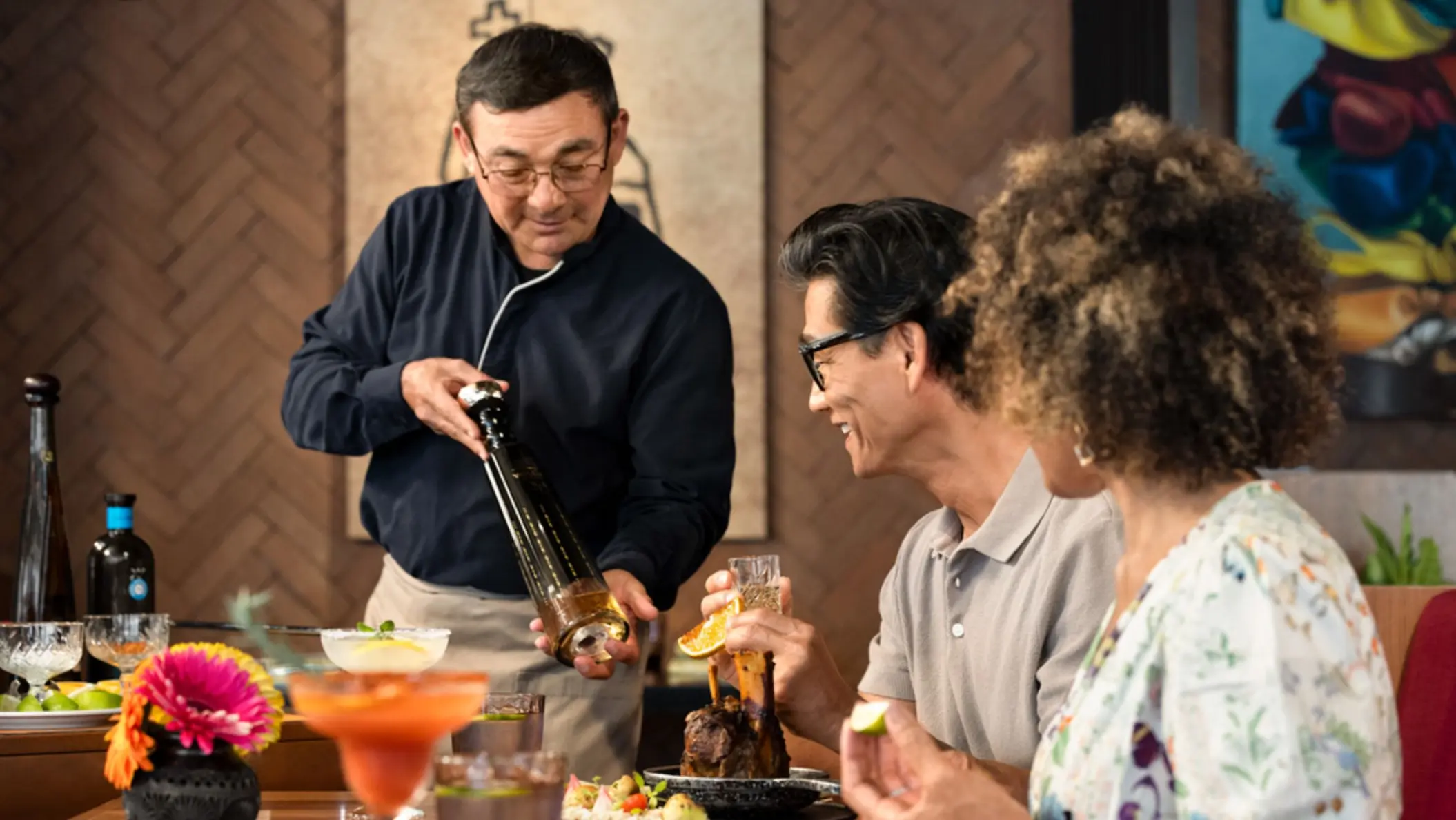 A bartender is pouring from a bottle for a smiling couple sitting at a table in a restaurant.