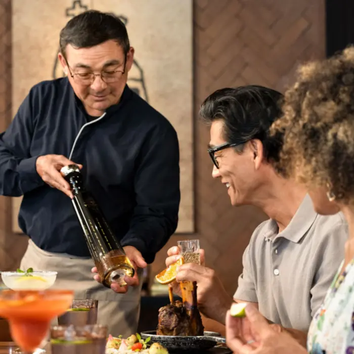 A bartender is pouring from a bottle for a smiling couple sitting at a table in a restaurant.