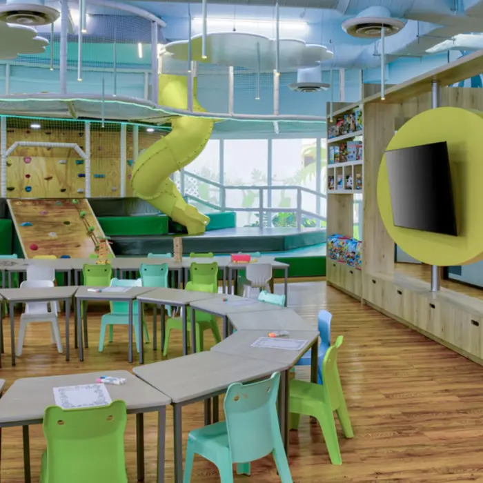 A bright and colorful indoor kids' play area with a yellow slide, climbing walls, and a round table with chairs for crafts.