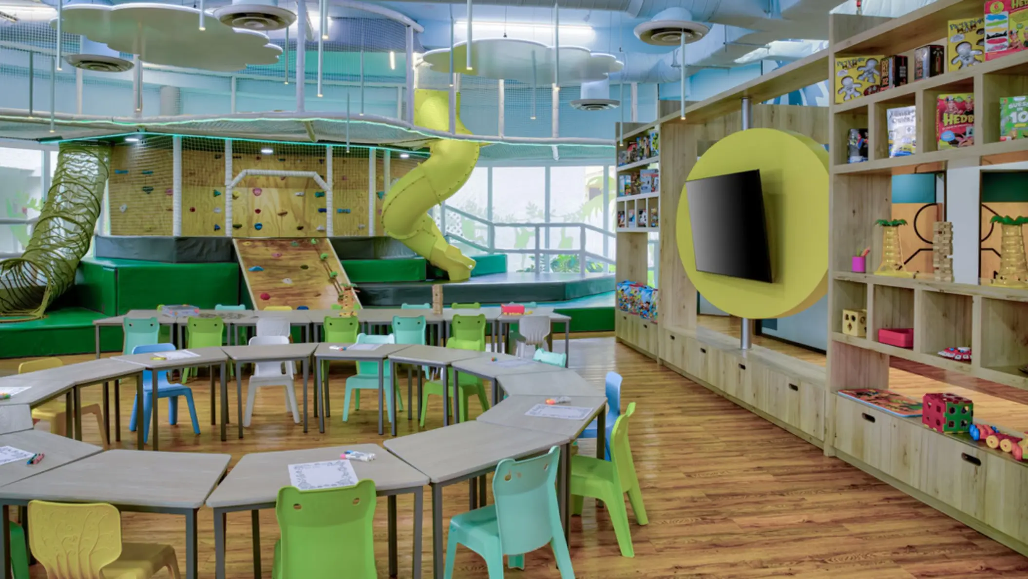 A bright and colorful indoor kids' play area with a yellow slide, climbing walls, and a round table with chairs for crafts.