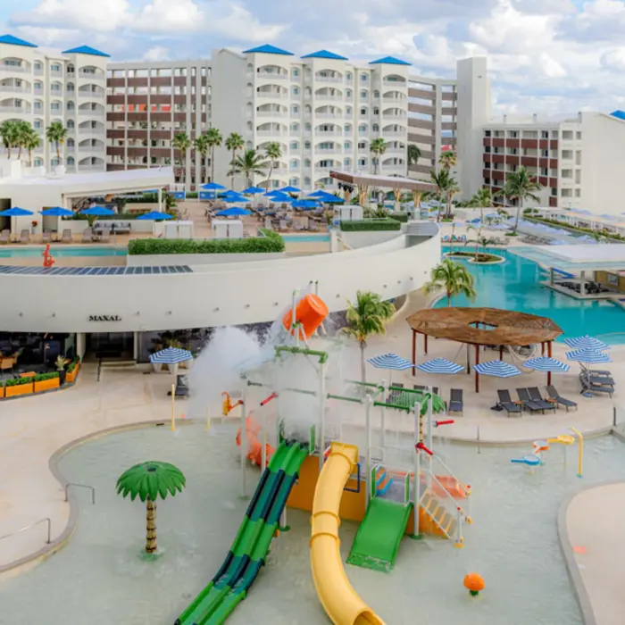 An aerial view of a vibrant hotel pool area with a kids' water park, slides, and lounge chairs surrounded by resort buildings.