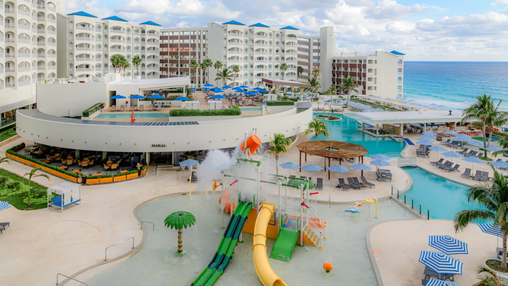 An aerial view of a vibrant hotel pool area with a kids' water park, slides, and lounge chairs surrounded by resort buildings.