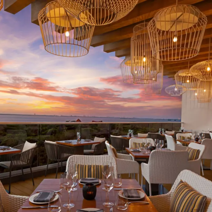 A stunning restaurant terrace overlooking a body of water at sunset, with wicker chairs and tables set for dining.