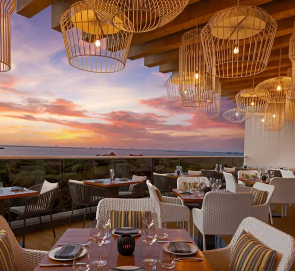 A stunning restaurant terrace overlooking a body of water at sunset, with wicker chairs and tables set for dining.