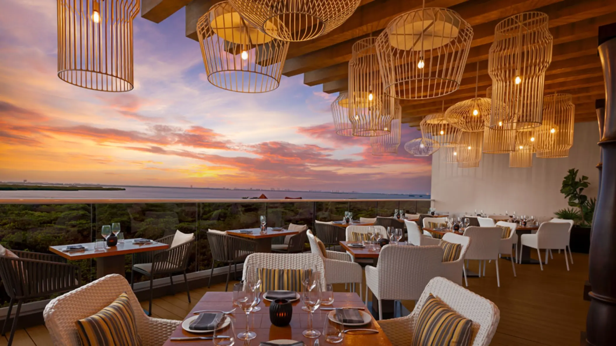 A stunning restaurant terrace overlooking a body of water at sunset, with wicker chairs and tables set for dining.