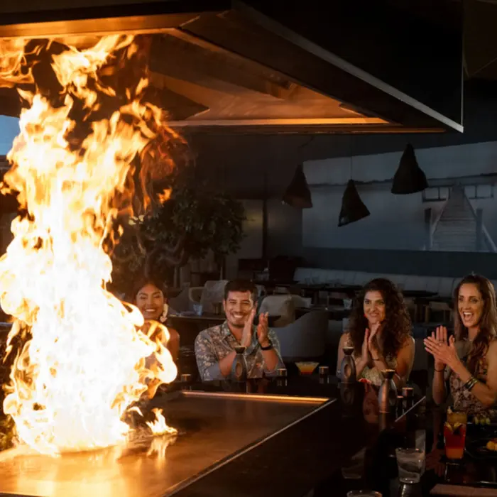 A teppanyaki chef creating a dramatic flame display, entertaining a group of smiling and applauding diners.