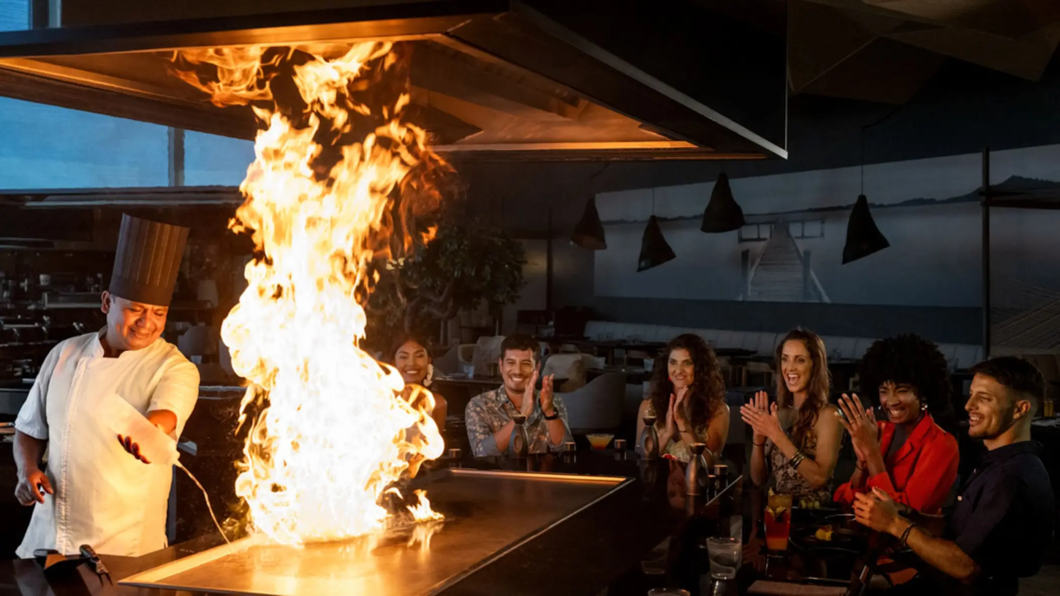 A teppanyaki chef creating a dramatic flame display, entertaining a group of smiling and applauding diners.