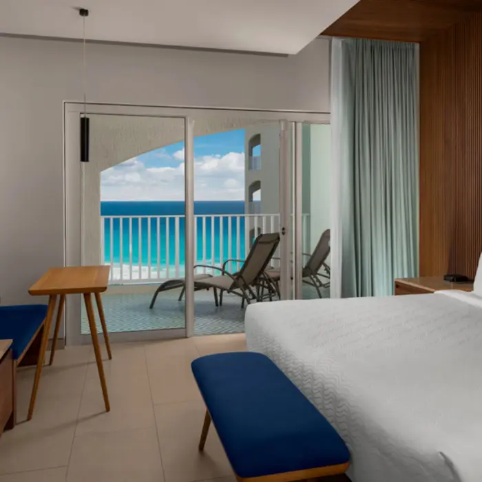 A stylish ocean-view hotel room with a modern bed, a small table, a TV, and a balcony with chairs.