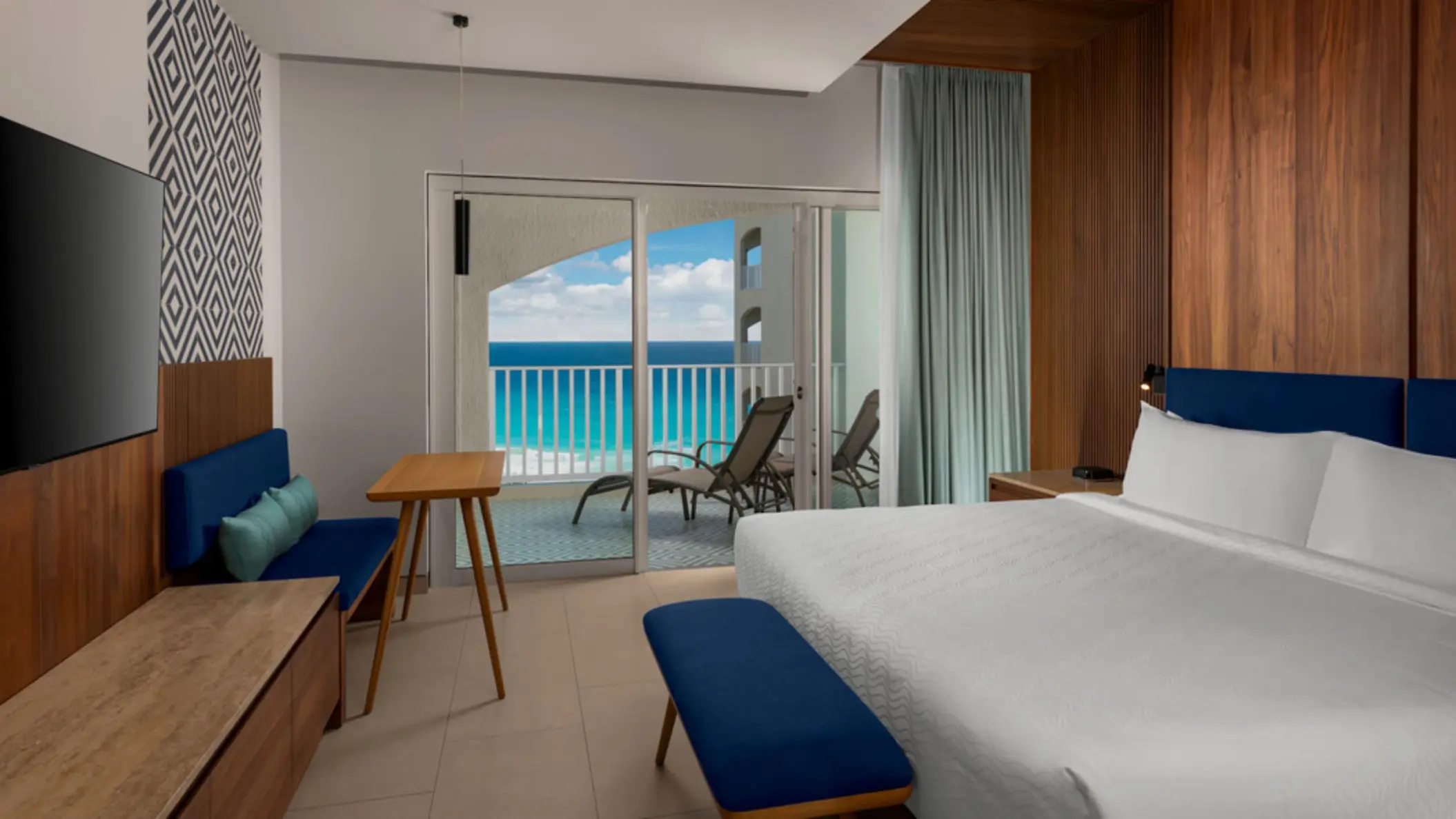 A stylish ocean-view hotel room with a modern bed, a small table, a TV, and a balcony with chairs.