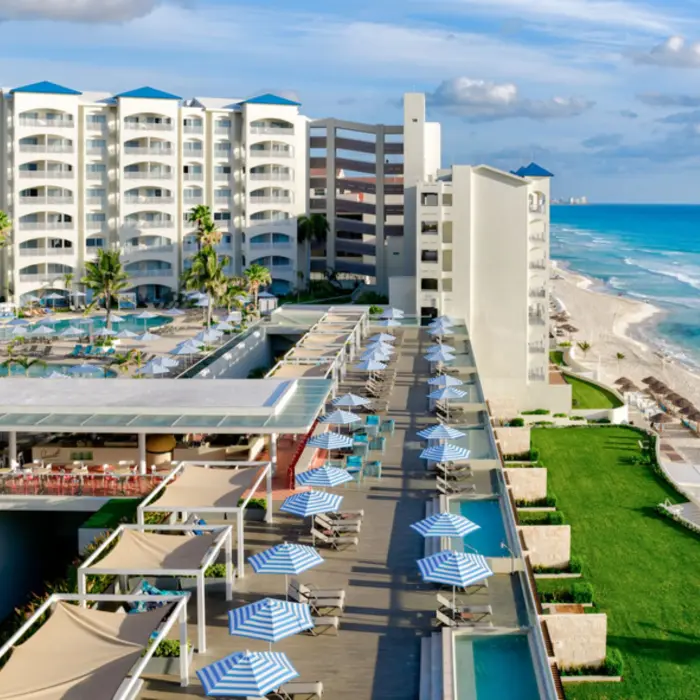 n aerial view of a large beach resort with swimming pools, a green lawn, and beach chairs lining the turquoise water.