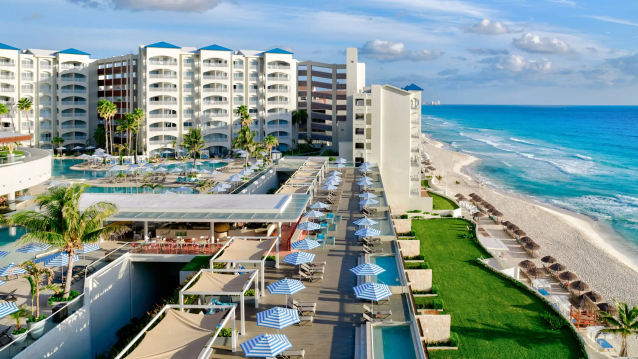 n aerial view of a large beach resort with swimming pools, a green lawn, and beach chairs lining the turquoise water.