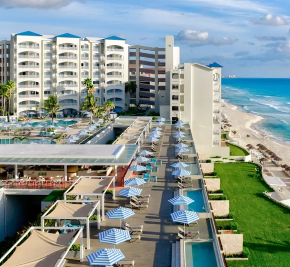 n aerial view of a large beach resort with swimming pools, a green lawn, and beach chairs lining the turquoise water.