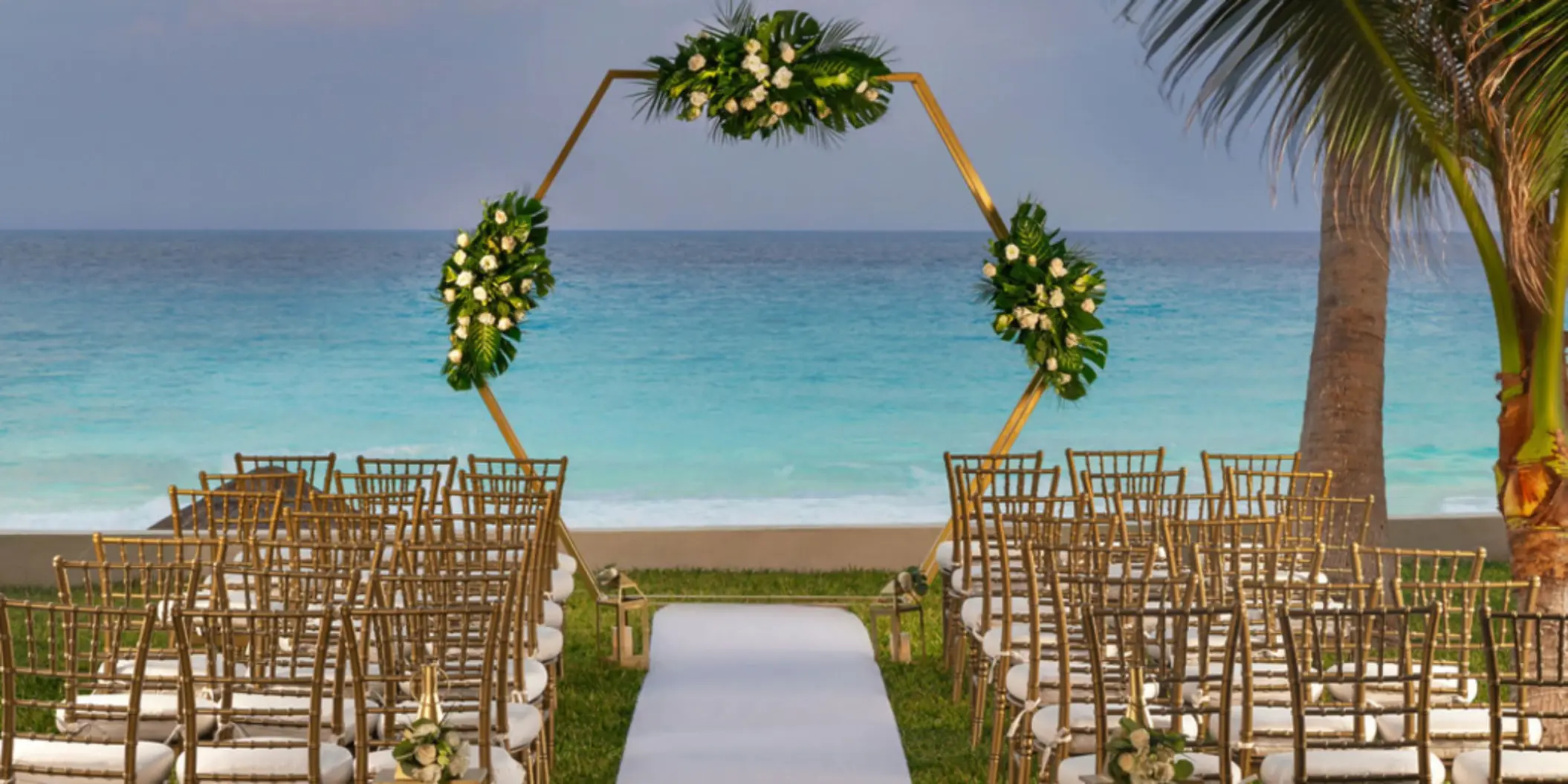 A stunning wedding venue with a golden geometric arch decorated with flowers, white chairs, and an ocean backdrop.