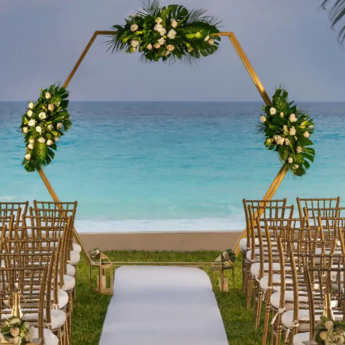 A stunning wedding venue with a golden geometric arch decorated with flowers, white chairs, and an ocean backdrop.