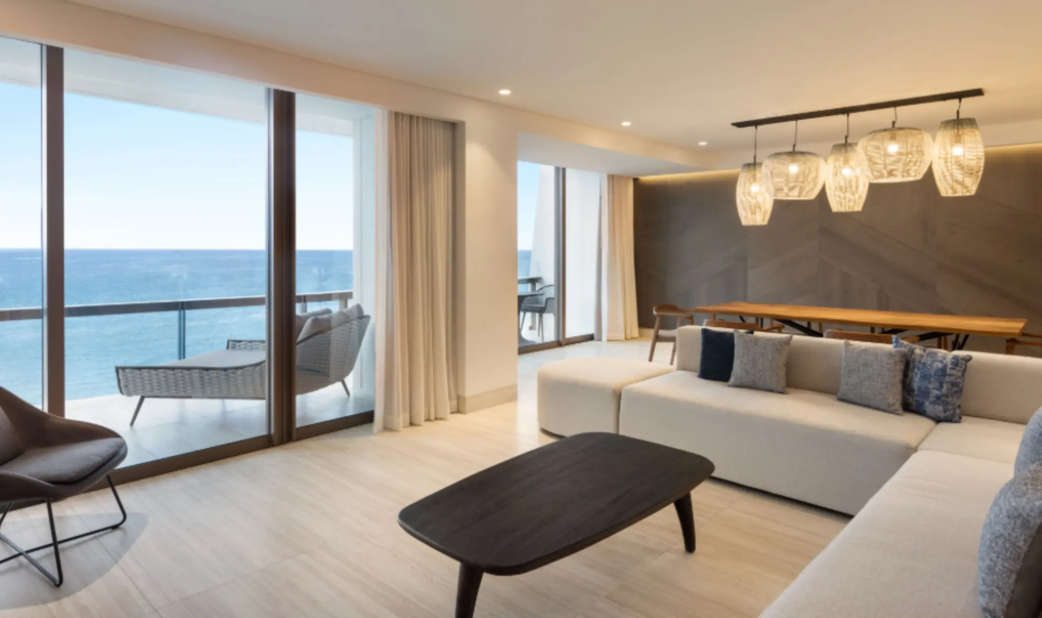 Oceanview suite with modern living area, large sectional, wooden dining table, and access to a private balcony.