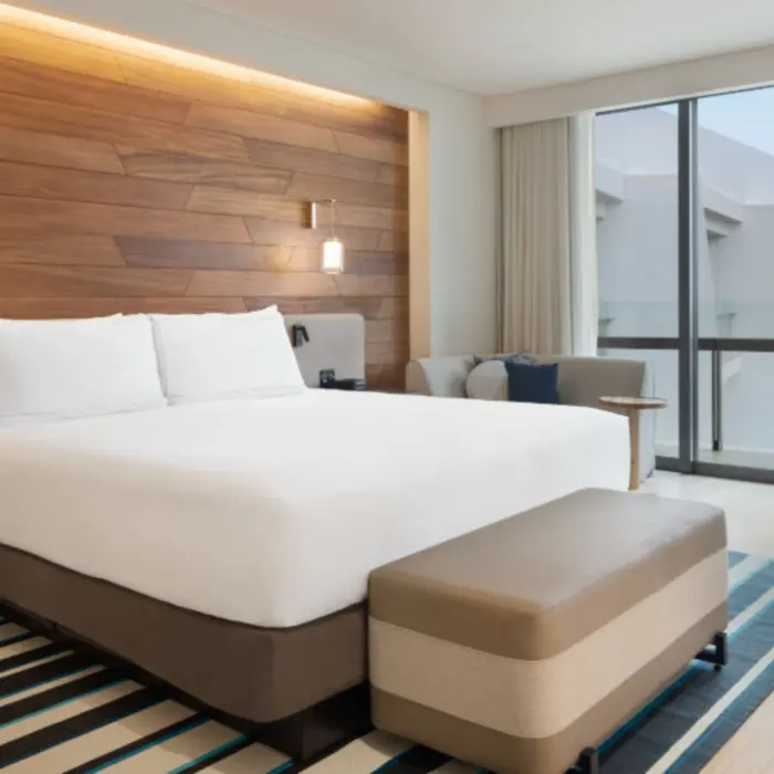 Contemporary guest room with king bed, wood-paneled headboard, cozy sitting area, and floor-to-ceiling windows.