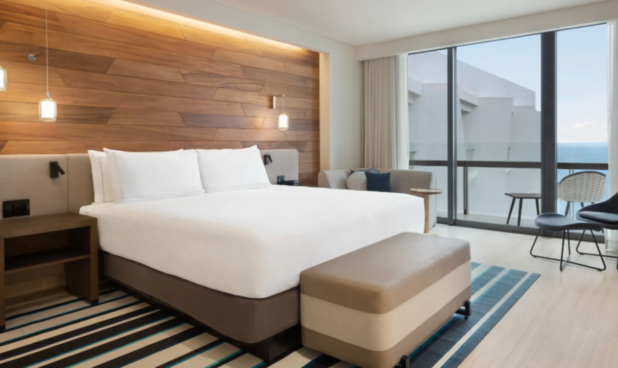 Contemporary guest room with king bed, wood-paneled headboard, cozy sitting area, and floor-to-ceiling windows.