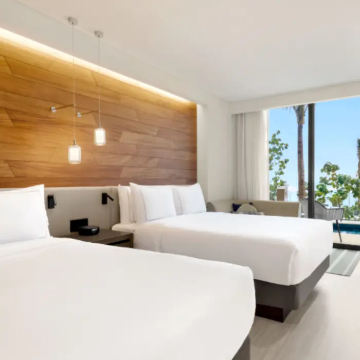 Bright room with two queen beds, wooden headboard, and a view of the plunge pool and lush greenery outside.