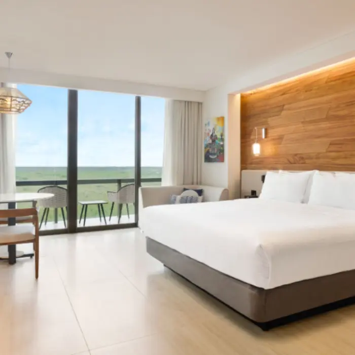 Stylish guest room with king bed, light wood floors, and private balcony overlooking tropical landscape.