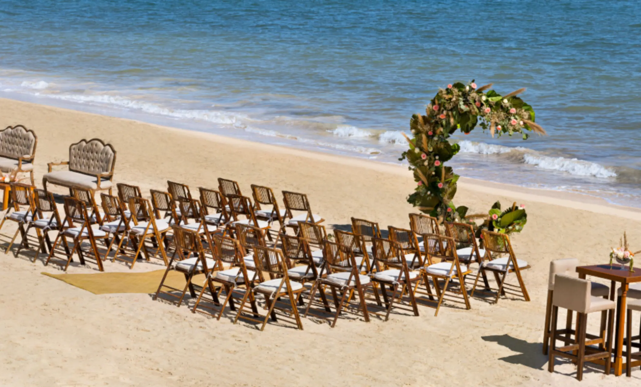 Romantic beach wedding setup with wooden chairs, tropical floral arch, and calm waves rolling onto the shore.