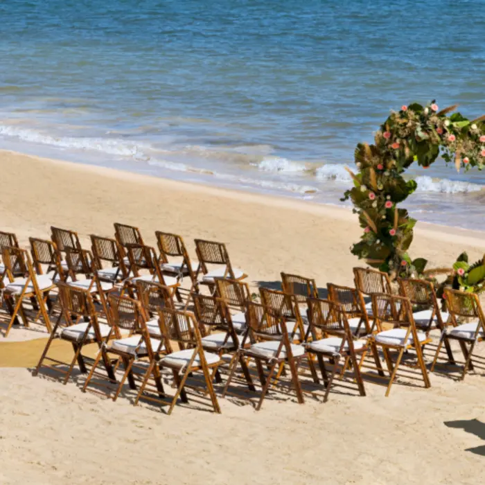 Romantic beach wedding setup with wooden chairs, tropical floral arch, and calm waves rolling onto the shore.