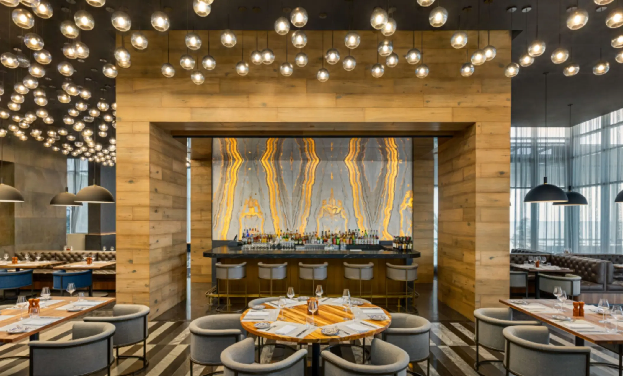 Elegant restaurant with glowing stone bar backdrop, wood paneling, and round tables set under globe pendant lights.