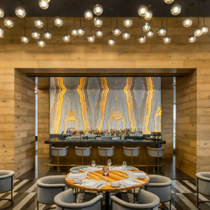 Elegant restaurant with glowing stone bar backdrop, wood paneling, and round tables set under globe pendant lights.