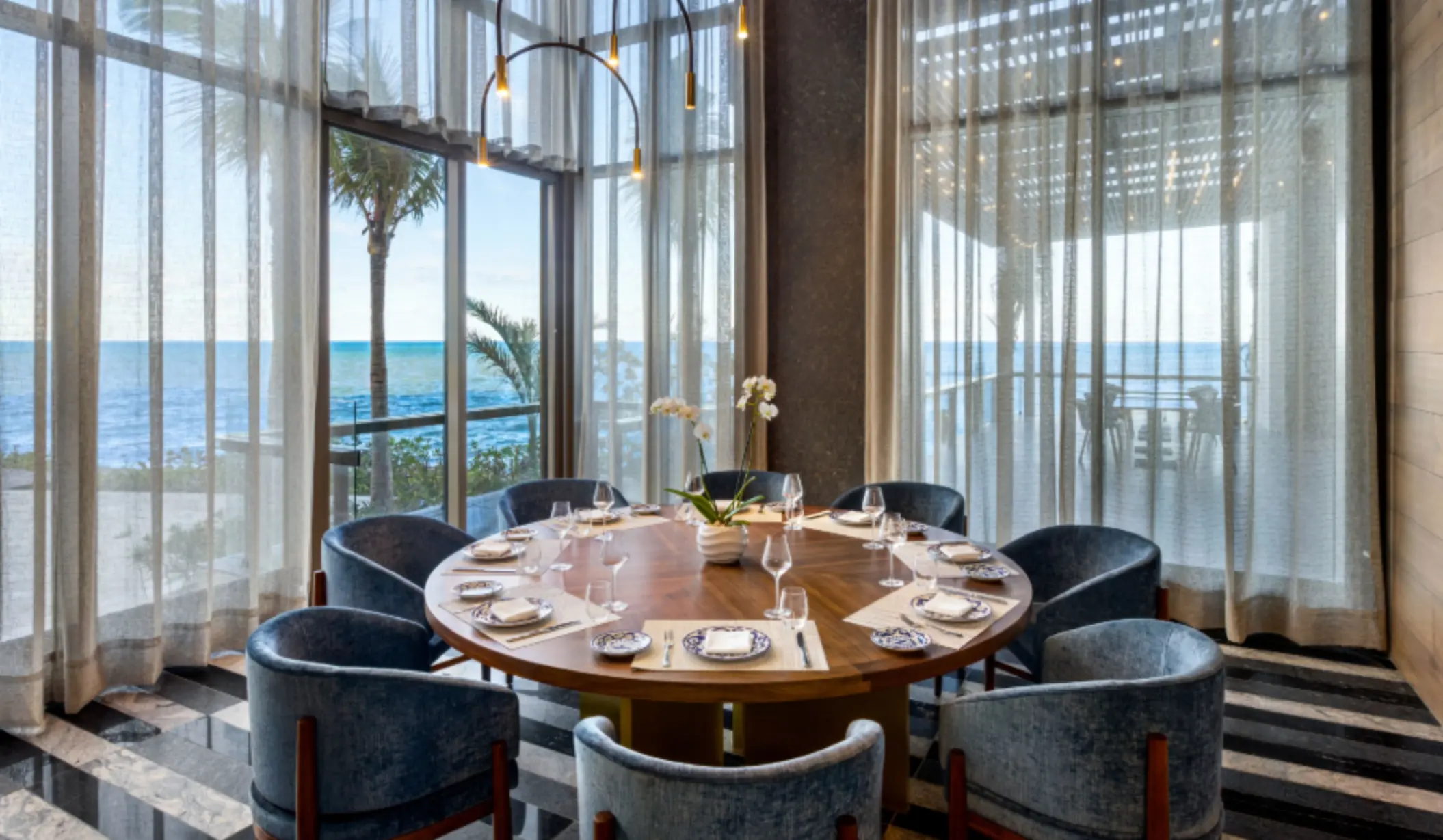Round dining table set by floor-to-ceiling windows with ocean views, velvet chairs, and natural light flooding the space.