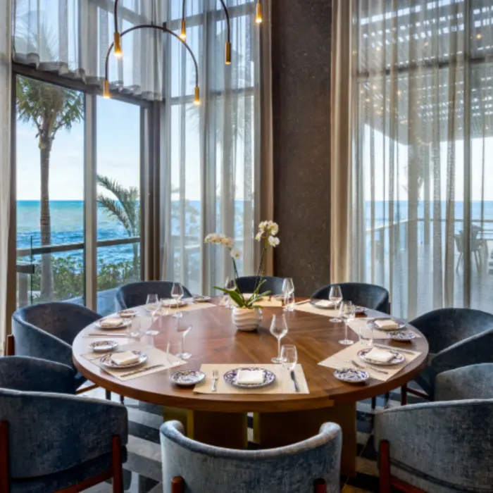 Round dining table set by floor-to-ceiling windows with ocean views, velvet chairs, and natural light flooding the space.
